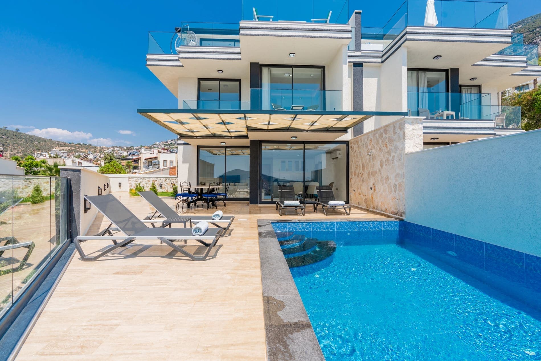 Gündüz saatlerinde havuz başında şezlonglar bulunan modern villa dış cephesi.