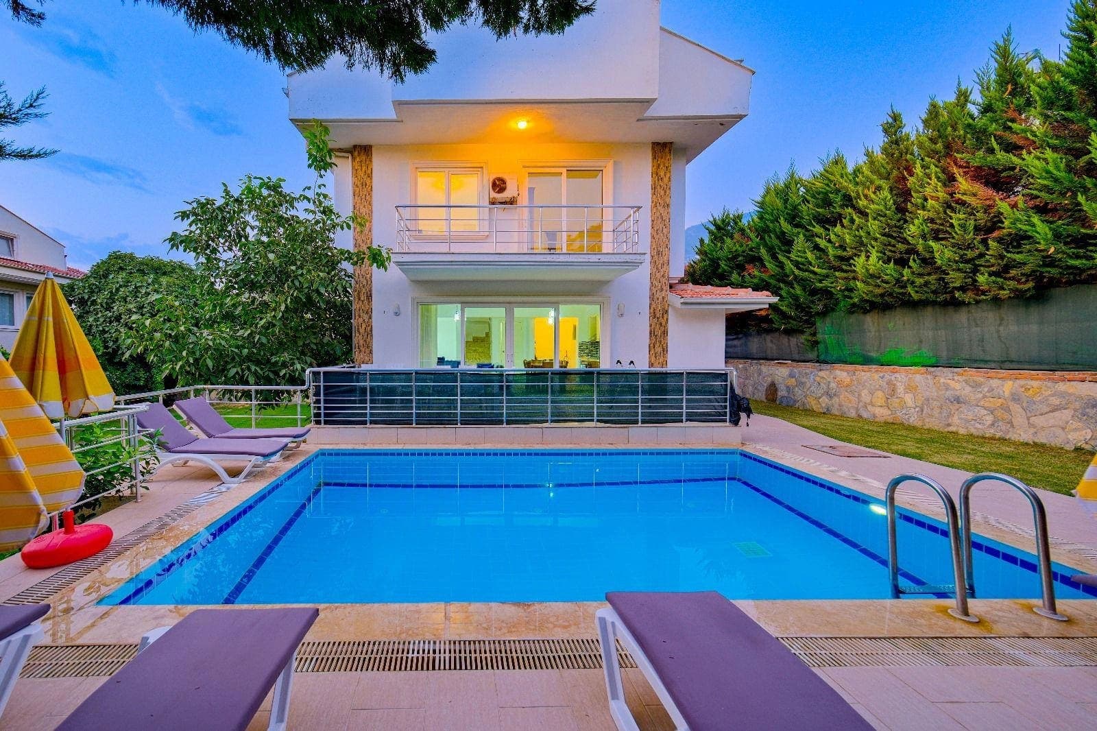 Akşam üstü ışıklandırılmış havuz ve önünde şezlonglar olan villa.
