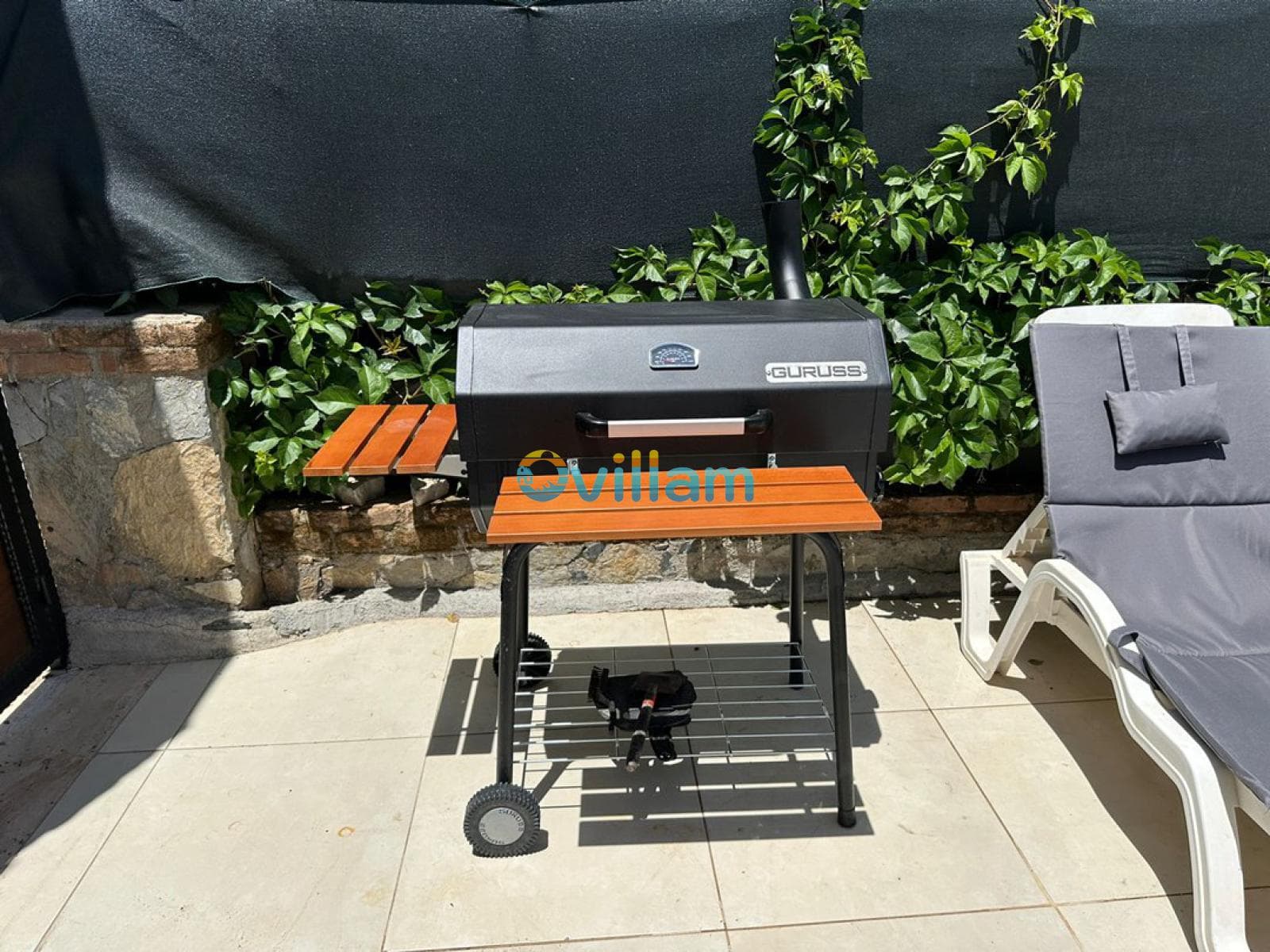 Teras zemininde siyah renkli tekerlekli barbekü ve yanında şezlong yer alıyor.