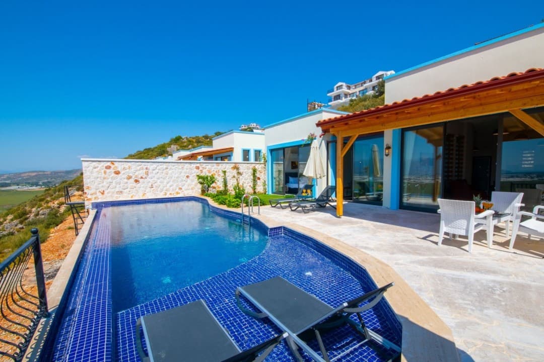 Mavi mozaik kaplı havuzu, şezlongları ve taş duvarlı terası olan, ahşap çatılı modern bir villa.