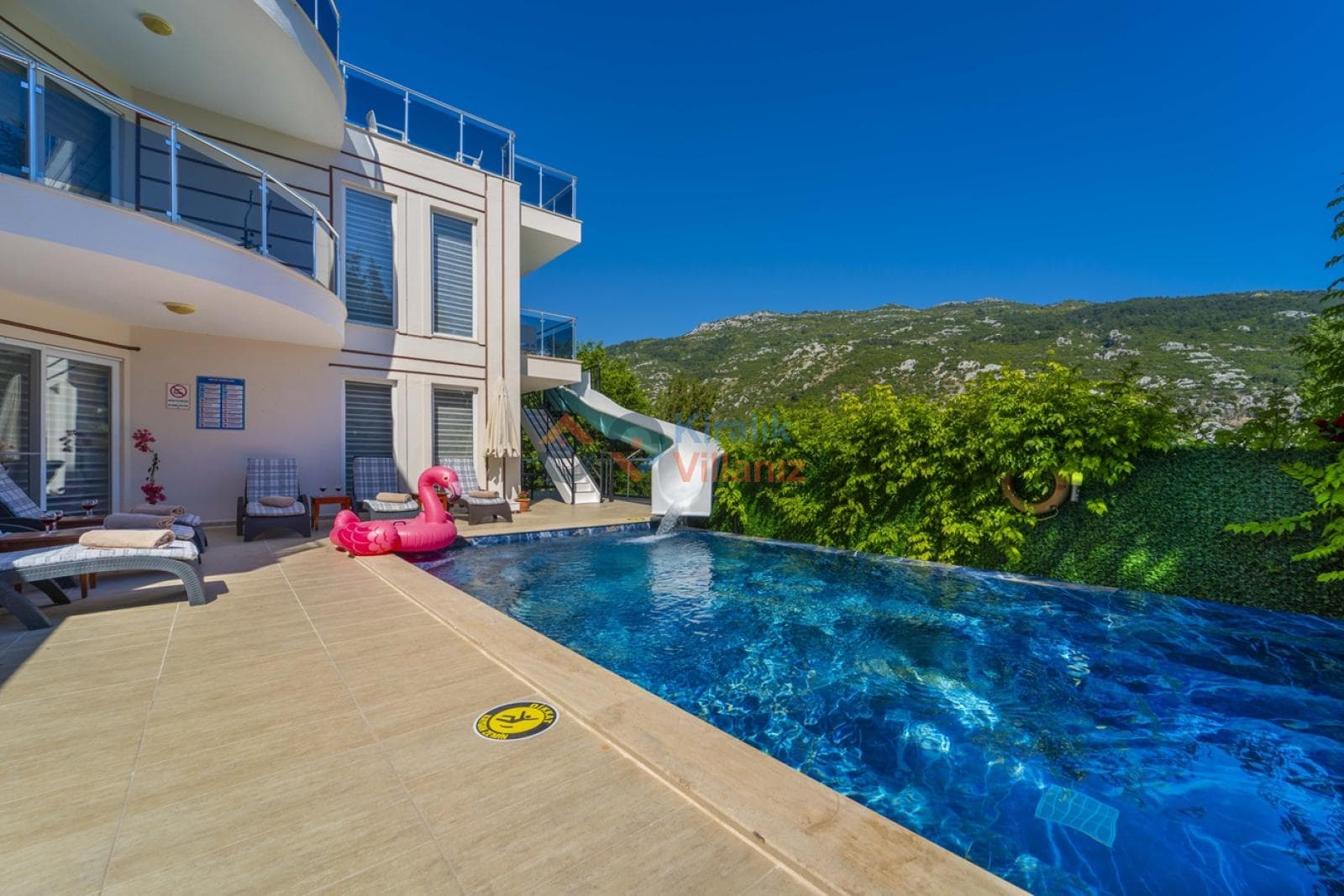 Güneşli günde havuz kenarında şezlonglar ve pembe flamingo simit bulunan villa dış cephesi.
