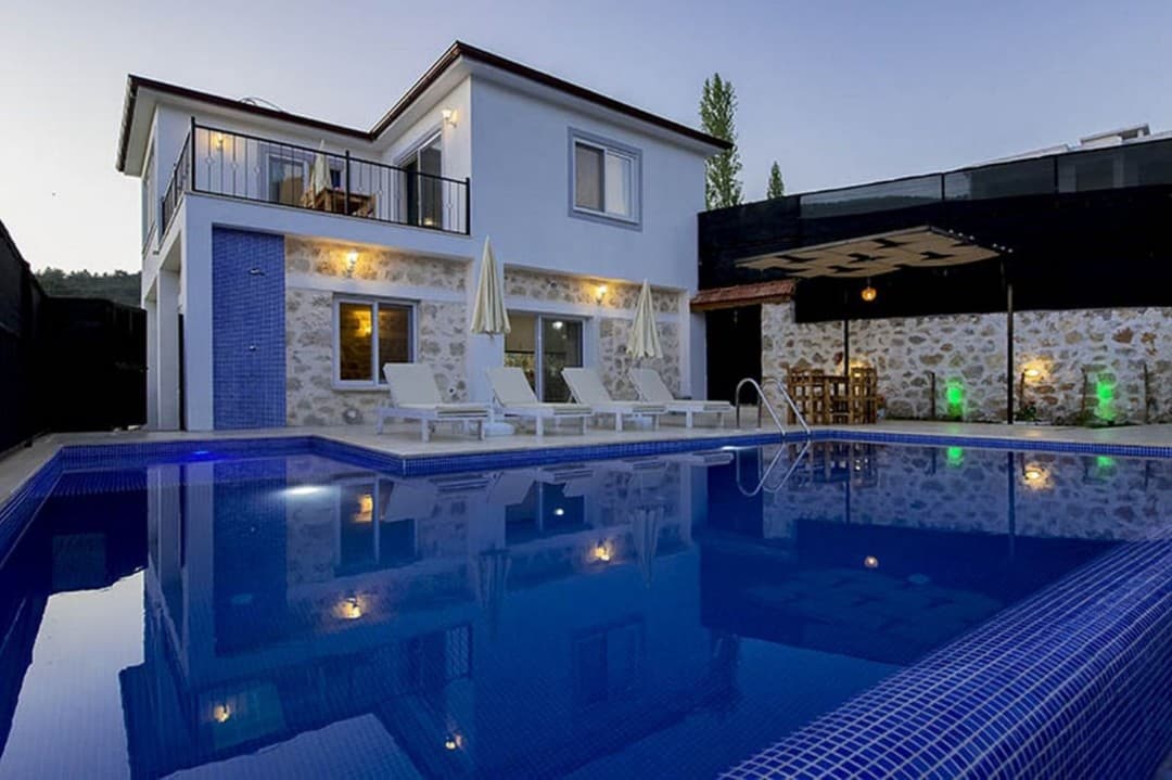 Akşam vakti aydınlatılmış havuzu ve şezlongları bulunan iki katlı beyaz villa.