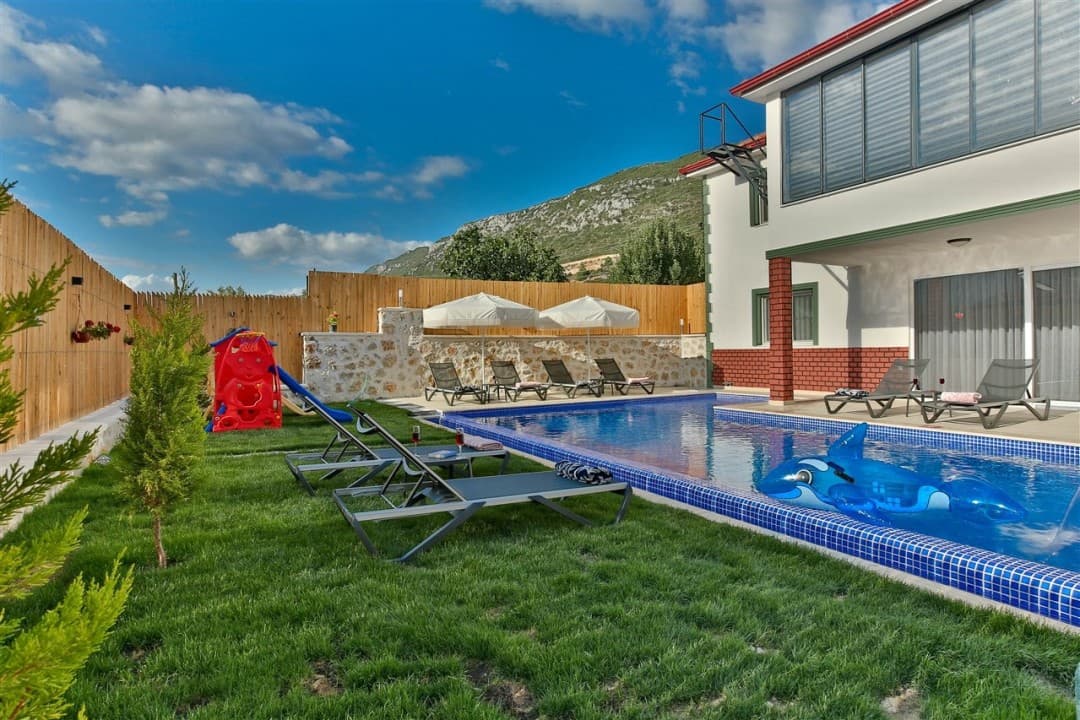 Havuz kenarında şezlonglar, çocuk kaydırağı ve arka planda dağ manzarası olan villa bahçesi.