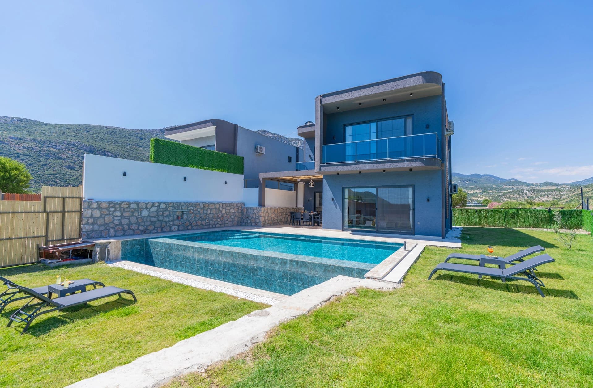 Önünde açık havuz ve şezlonglar bulunan, dağ manzaralı modern villa dış cephesi.