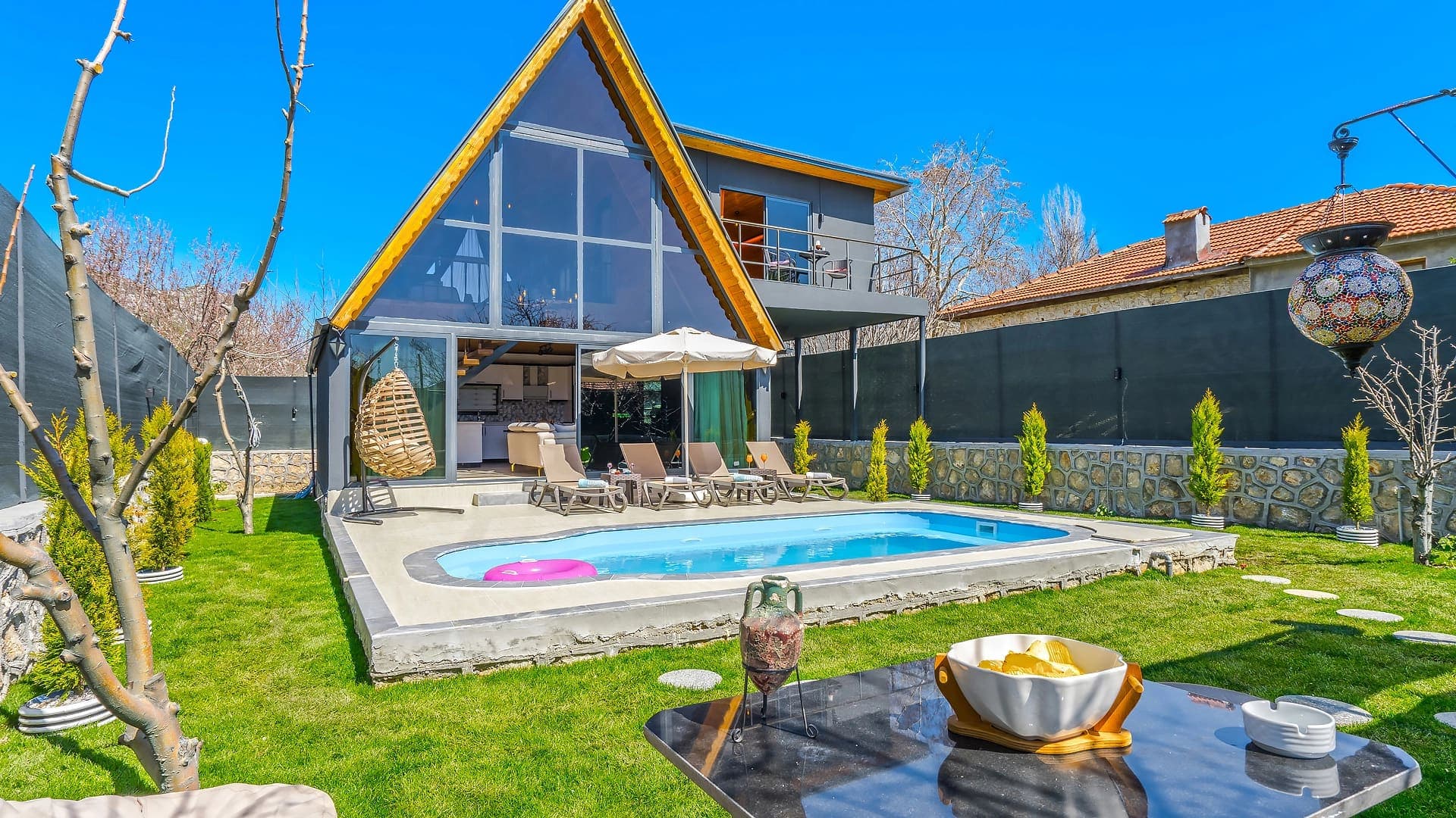Üçgen çatılı, cam cepheli ve önünde havuz bulunan villa bahçesi.