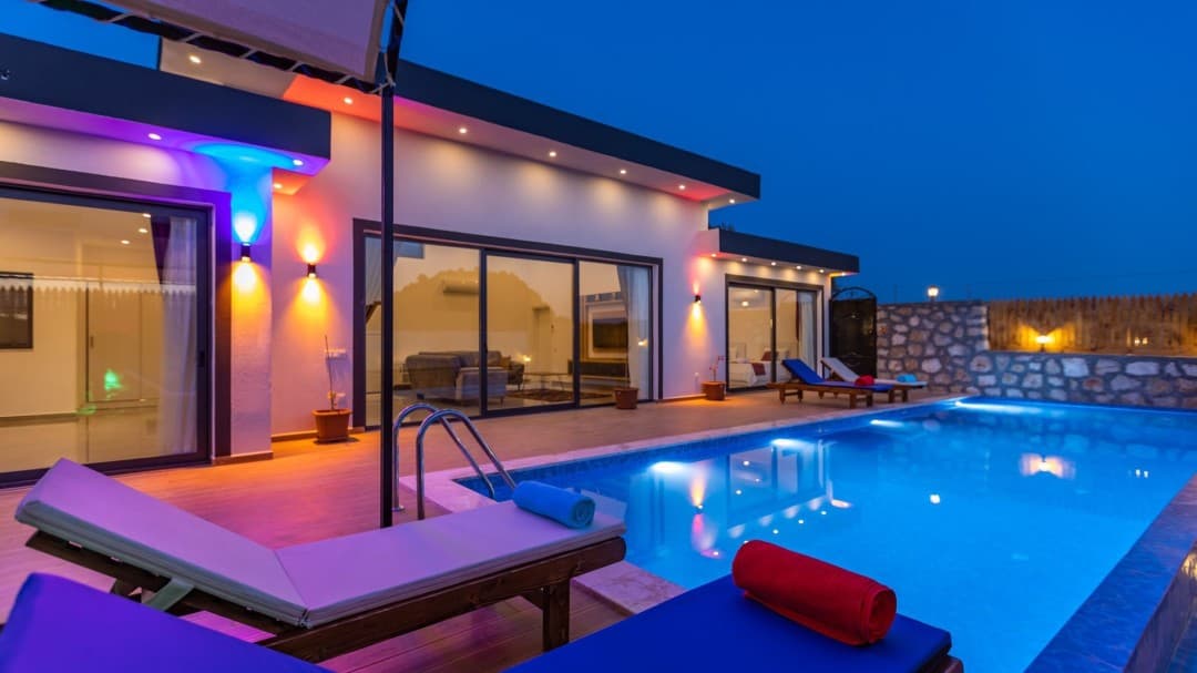 Gece aydınlatmalı, havuz kenarında şezlonglar bulunan modern villanın dış cephesi.