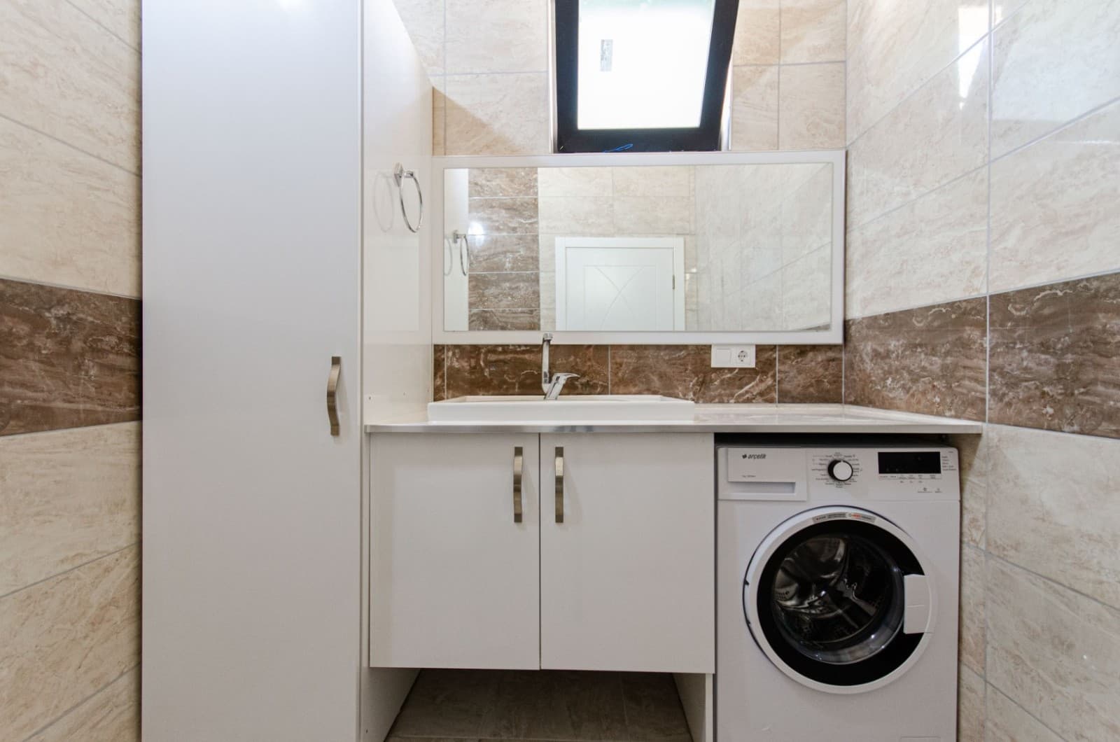 Bej seramik kaplı banyoda beyaz dolap, lavabo, çamaşır makinesi ve üstte pencere.