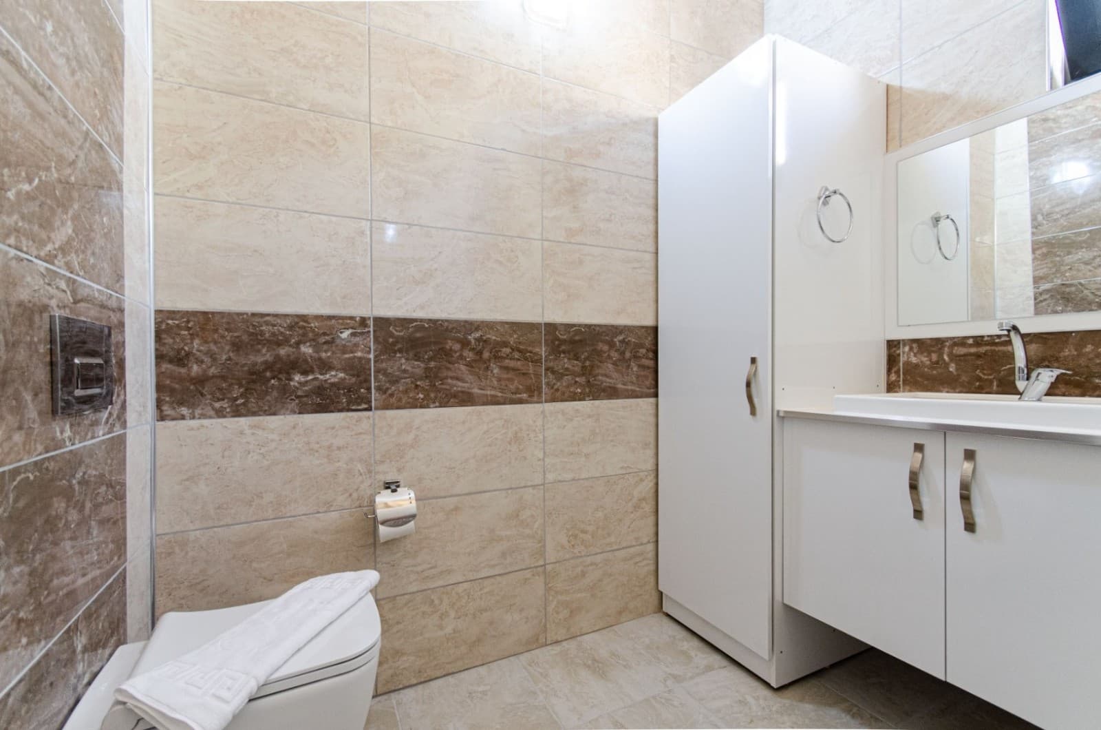 Bej ve kahverengi seramik kaplı banyoda beyaz lavabo dolabı ve asma klozet.