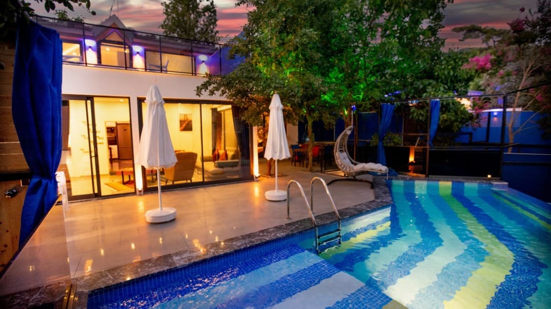 Akşam saatlerinde aydınlatmalı havuz ve iki şemsiyenin yer aldığı villa bahçesi.