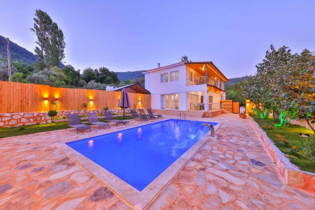 Akşam ışığında havuzlu, taş zeminli bahçesi ve ahşap çitleri olan iki katlı beyaz bir villa.