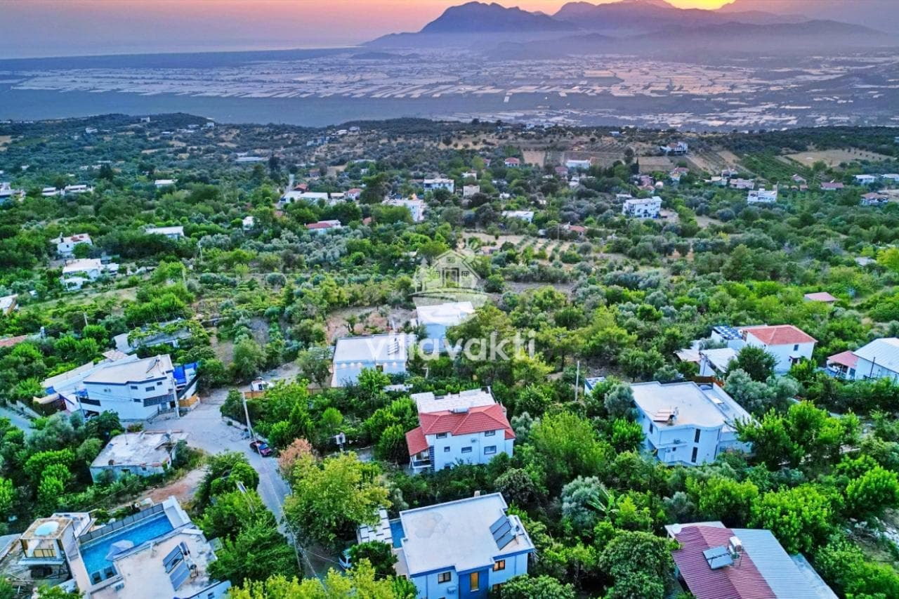 Ağaçlarla kaplı tepelerin ve uzakta deniz manzarasının göründüğü yüksek açıdan genel görünüm.