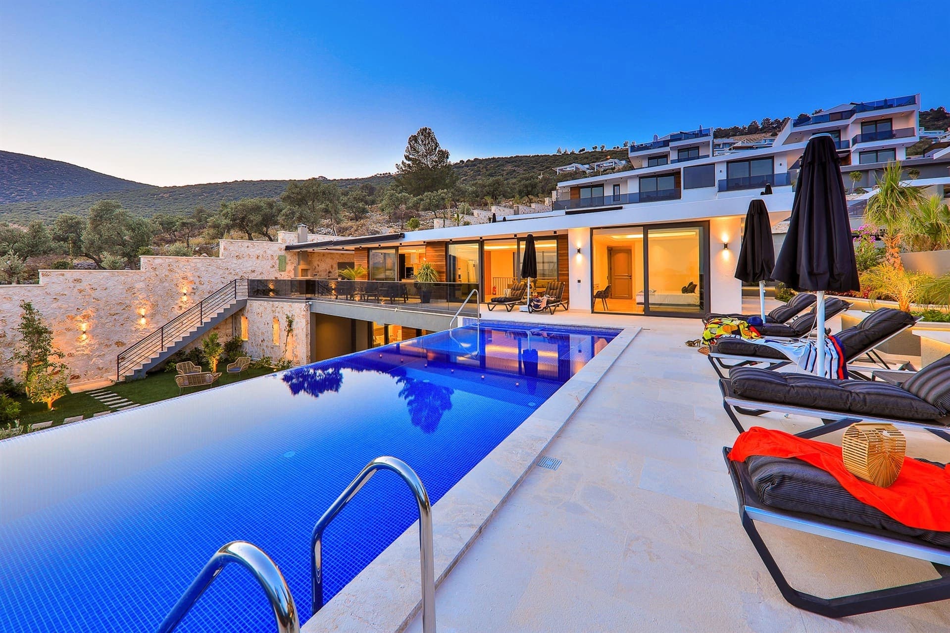 Aydınlatmalı havuz kenarında şezlonglar ve arka planda modern villa.