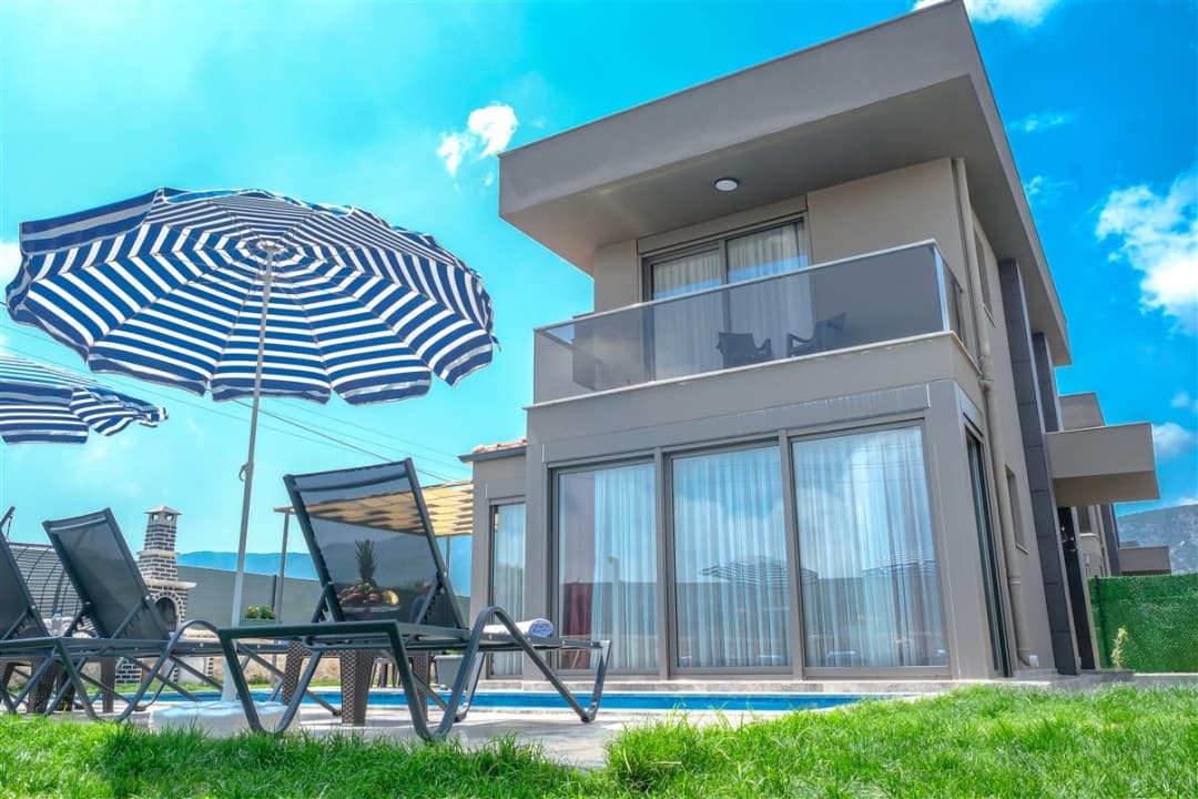 Mavi beyaz çizgili şemsiye, şezlonglar ve havuz kenarındaki modern villa dış cephesi.