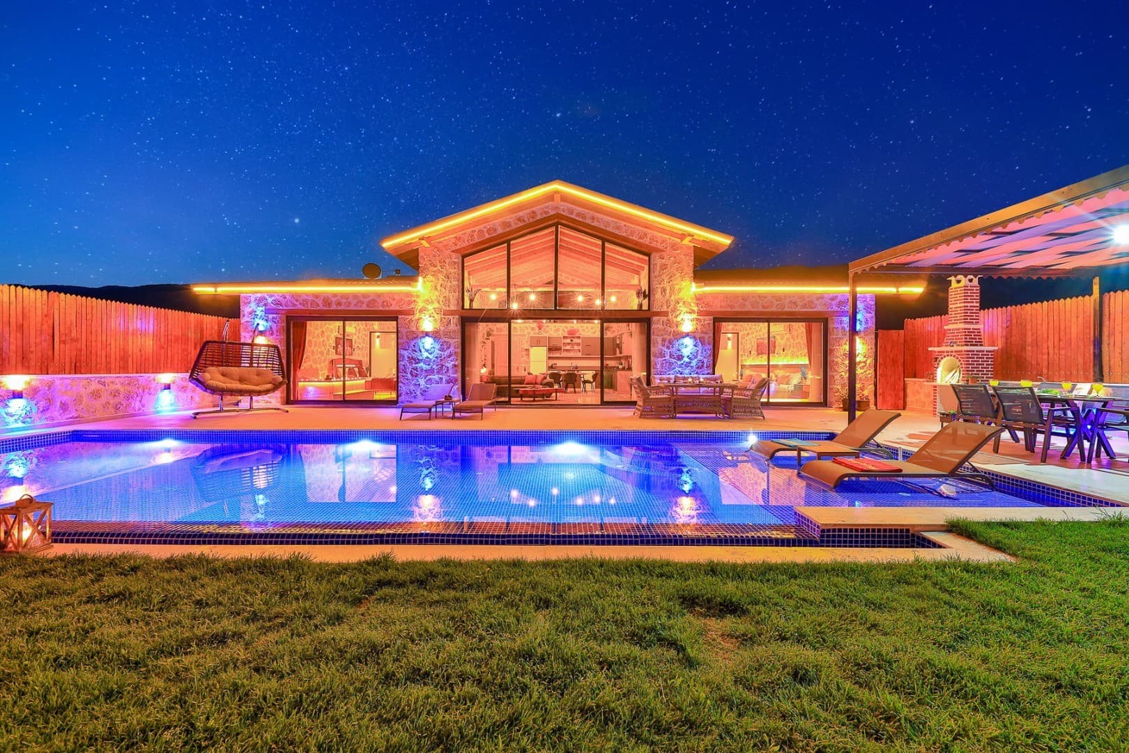 Aydınlatmalı havuzu, şezlongları ve yıldızlı gökyüzü altında taş duvarlı villa.