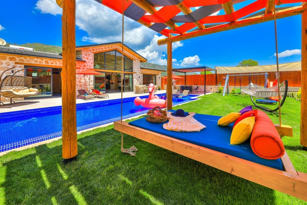 Bahçede asılı yatak, mavi yüzme havuzu, flamingo deniz yatağı ve taş duvarlı villa binası.