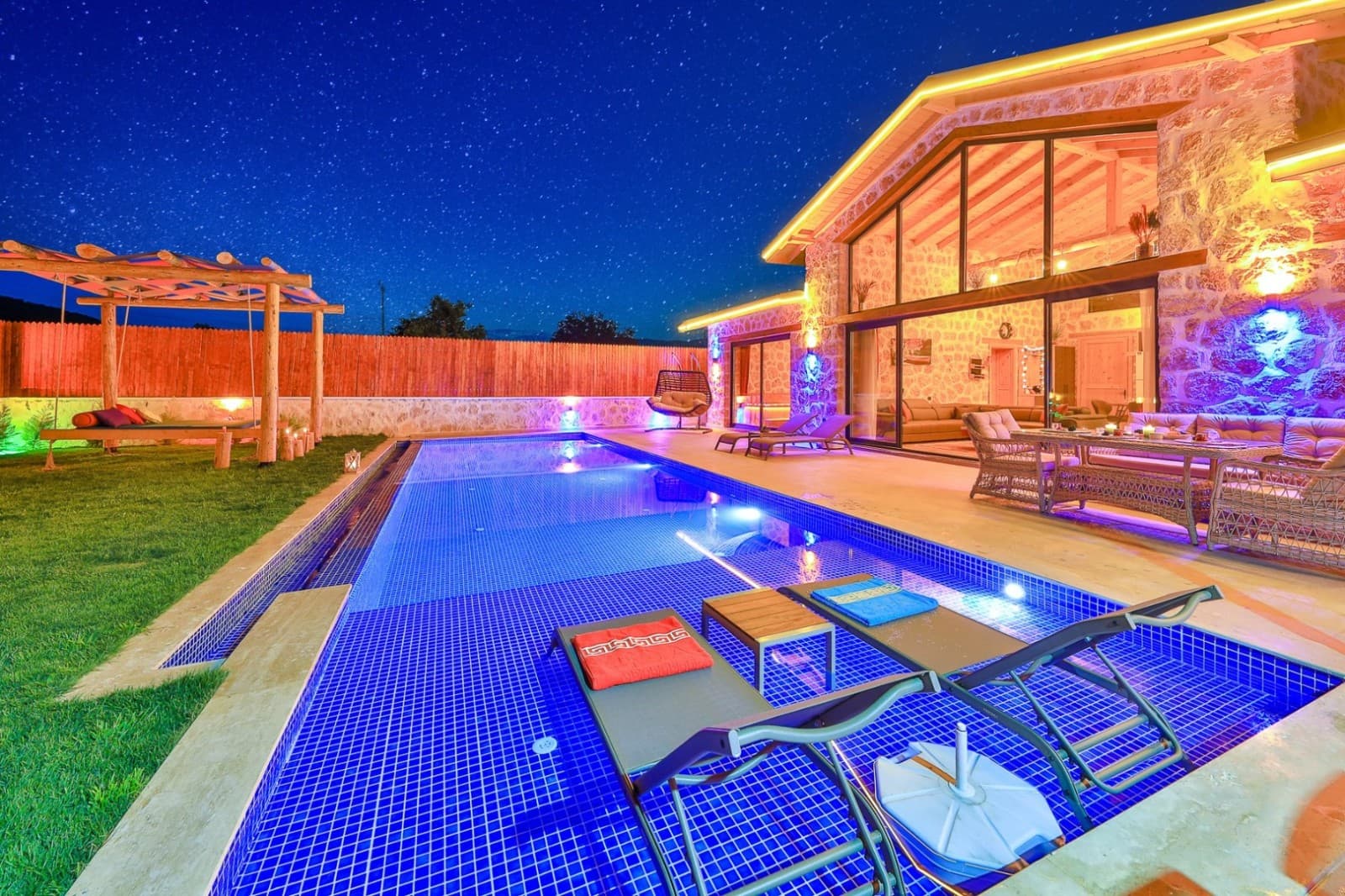 Gece aydınlatılmış taş villa, havuz, şezlonglar ve yıldızlı gökyüzü.
