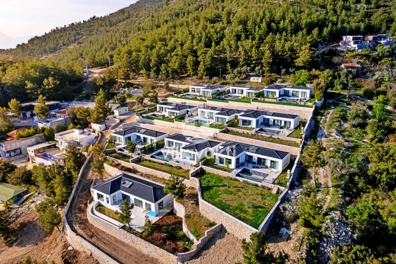 Ormanlık dağ yamacında kademeli olarak yerleşmiş havuzlu villaların havadan görünümü.