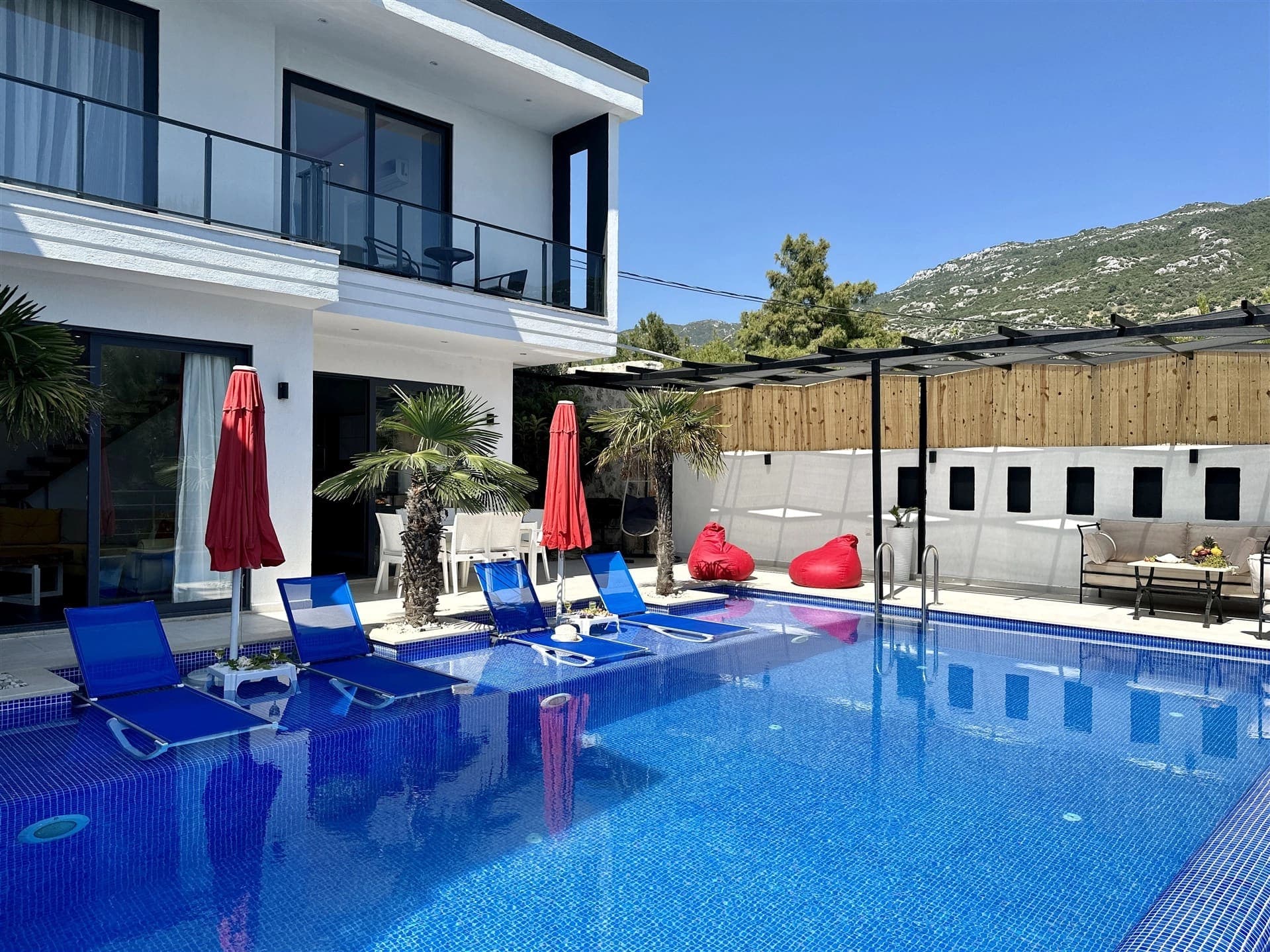 Mavi şezlonglu havuz ve arka planda dağ manzaralı beyaz villa dış cephesi.