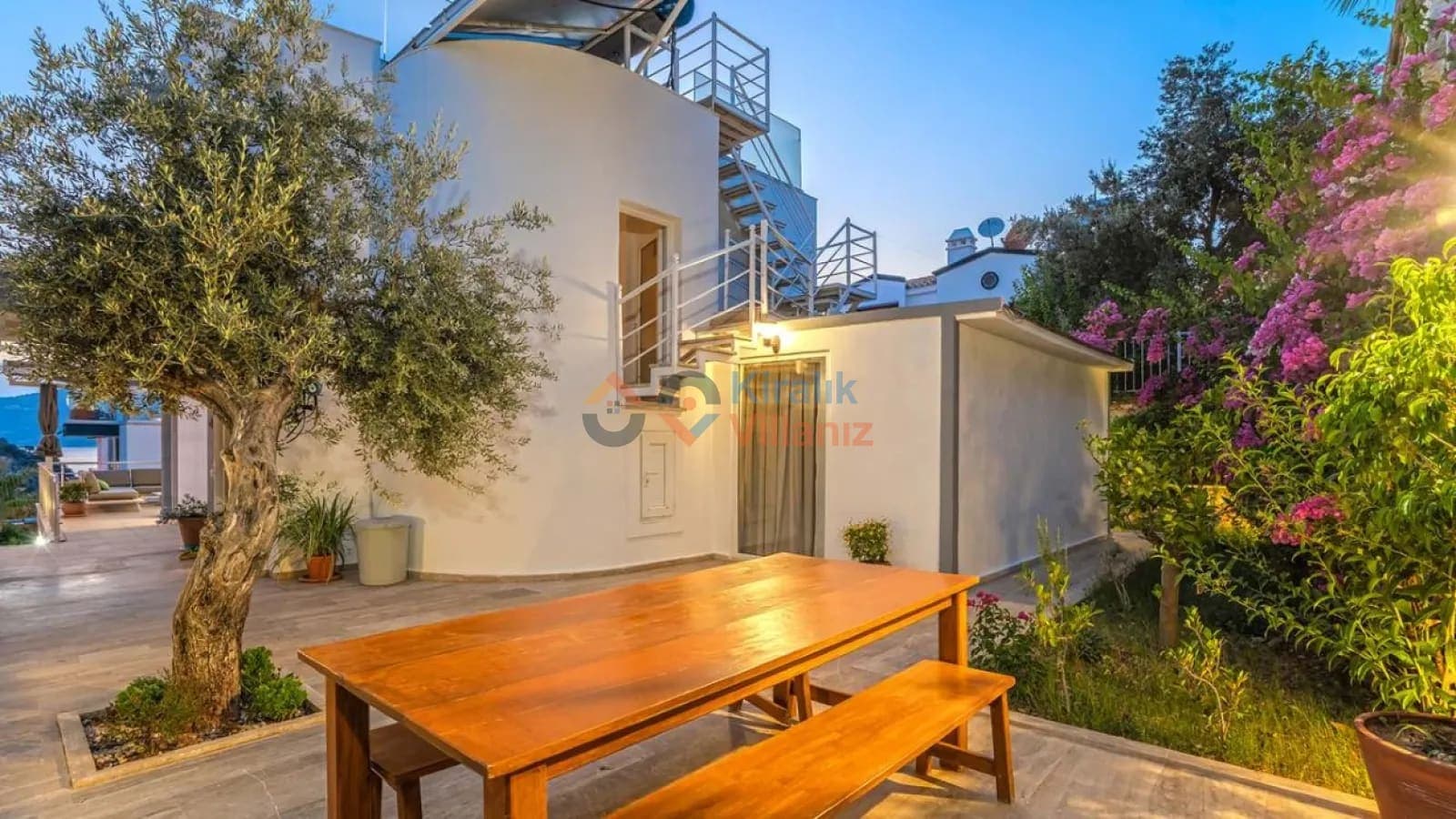 Bahçede ahşap yemek masası ve zeytin ağaçları bulunan villa dış cephesi.