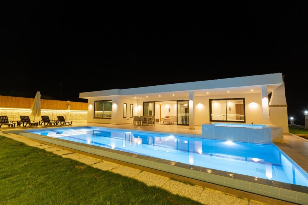 Gece vakti ışıklandırılmış havuzu ve şezlongları olan tek katlı beyaz villa.