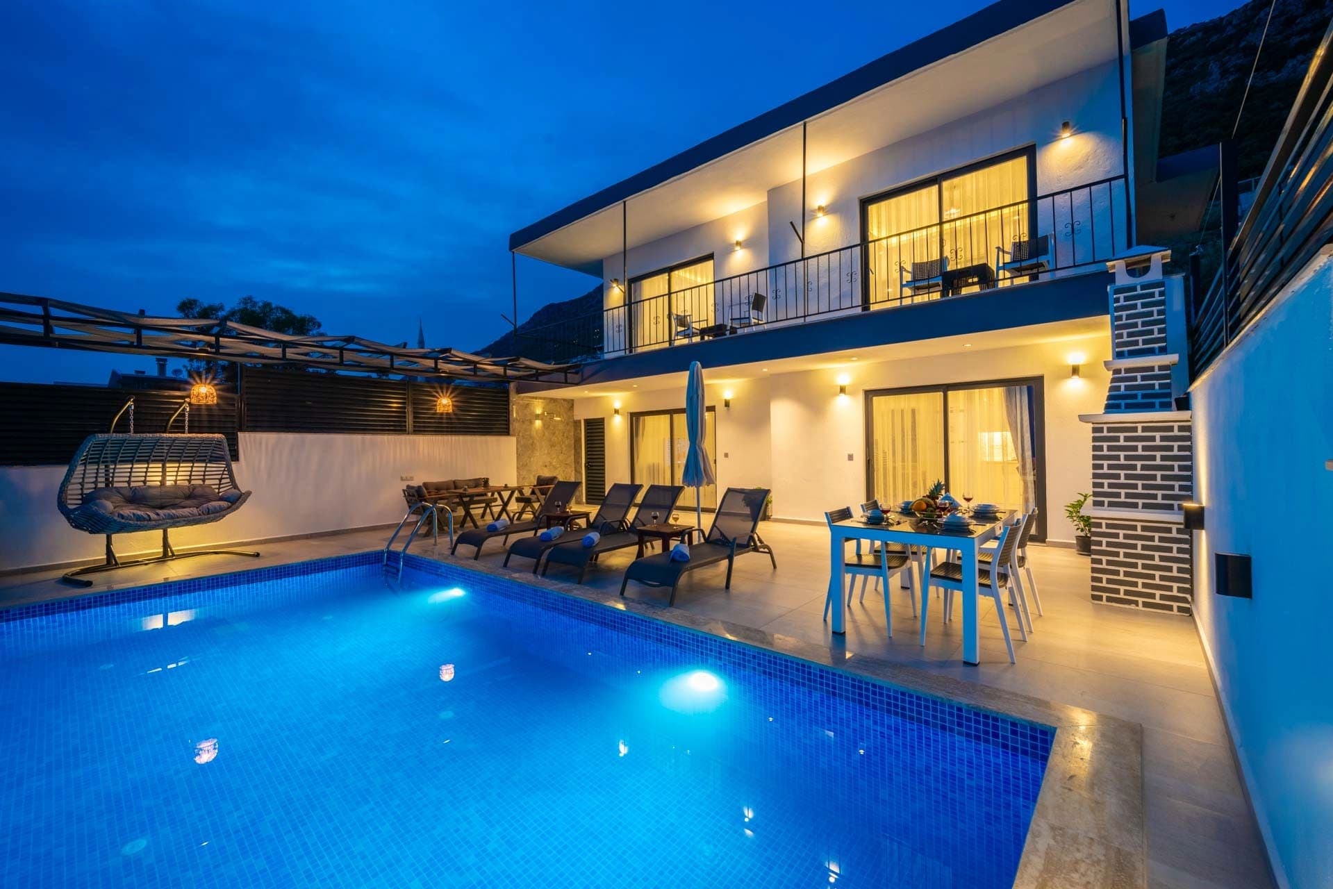 Aydınlatmalı havuz kenarında masa, sandalyeler ve iki katlı villanın gece görünümü.