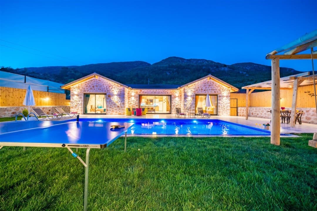 Akşam vakti aydınlatmalı taş villanın önünde havuz ve masa tenisi masası var.