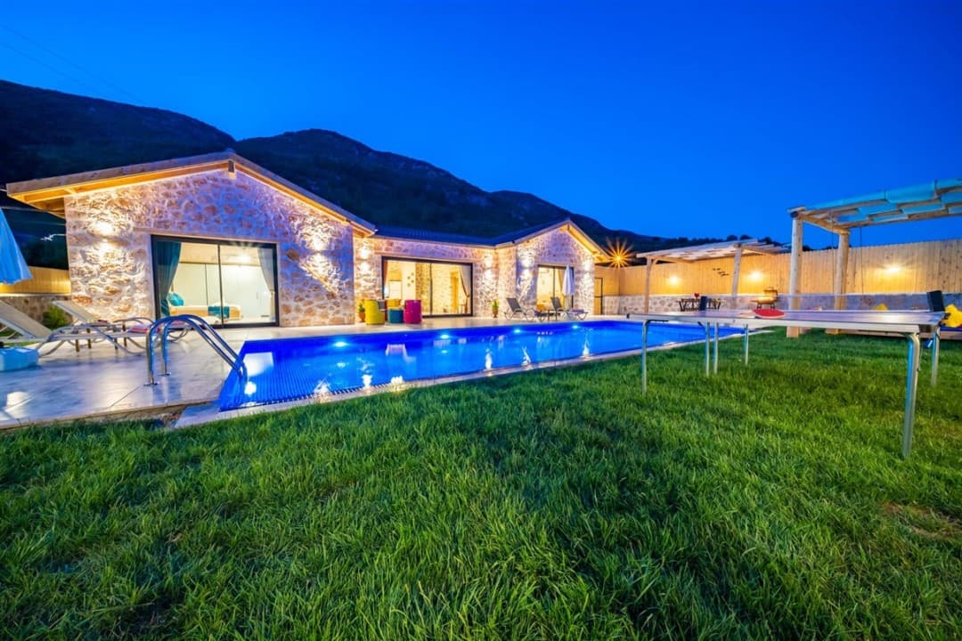 Gece aydınlatmalı taş villa, önünde yüzme havuzu, çim bahçe ve masa tenisi bulunan dış görünüm.
