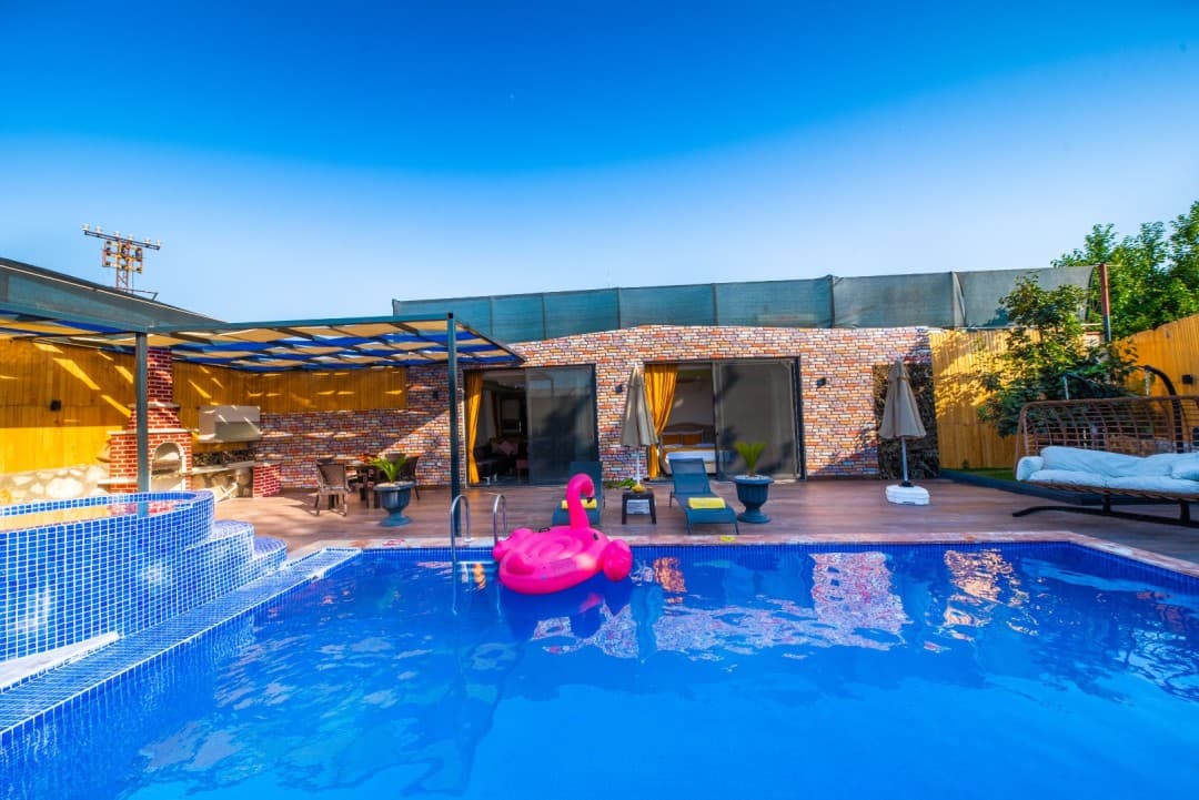 Önünde pembe flamingo deniz yatağı olan havuzlu, tuğla duvarlı ve ahşap zeminli villa bahçesi.