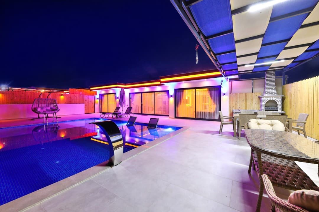 Gece aydınlatmalı havuz, şezlonglar, oturma grubu ve taş barbekü bulunan villanın dış mekânı.