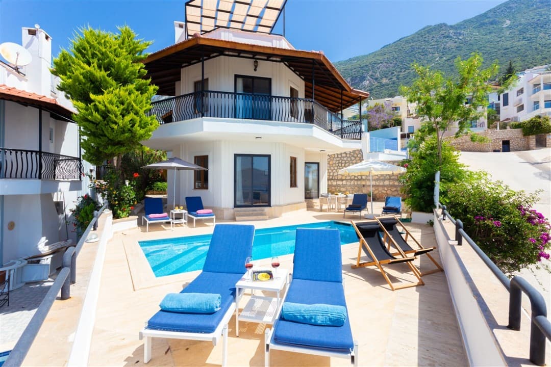 Mavi şezlonglu, havuzlu ve balkonlu beyaz dış cepheli iki katlı villa.
