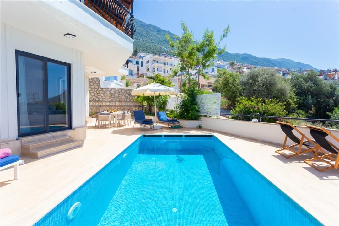 Mavi havuzlu, şezlonglu, güneş şemsiyeli ve dağ manzaralı villa terası.