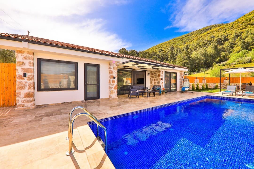 Mavi havuzlu, şezlong ve oturma alanı bulunan taş detaylı villa ile orman manzarası.