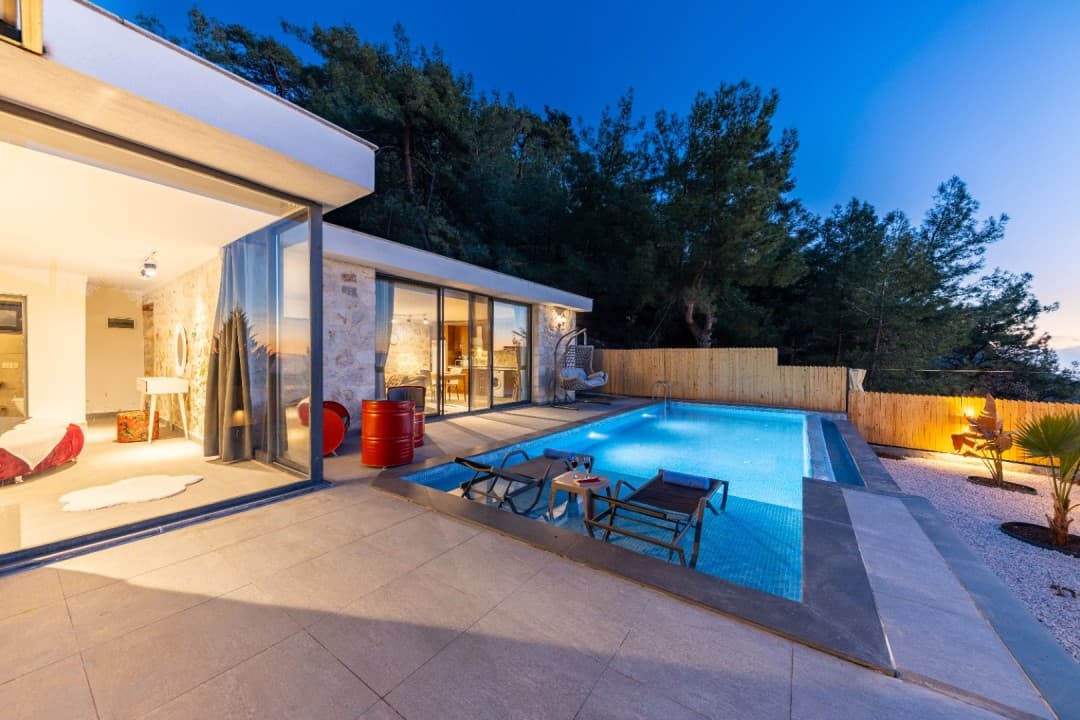 Akşam vakti aydınlatmalı yüzme havuzu, şezlonglar ve taş duvarlı modern villanın dış görünümü.