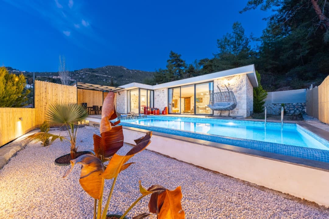 Akşam vakti ışıklandırılmış havuz, taş duvarlı villa, bahçe salıncağı ve dağ manzaralı çakıllı alan.