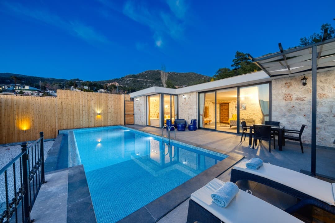 Akşam vakti aydınlatılmış havuzu, şezlongları ve taş duvarlı terası olan modern villa dış görünümü.