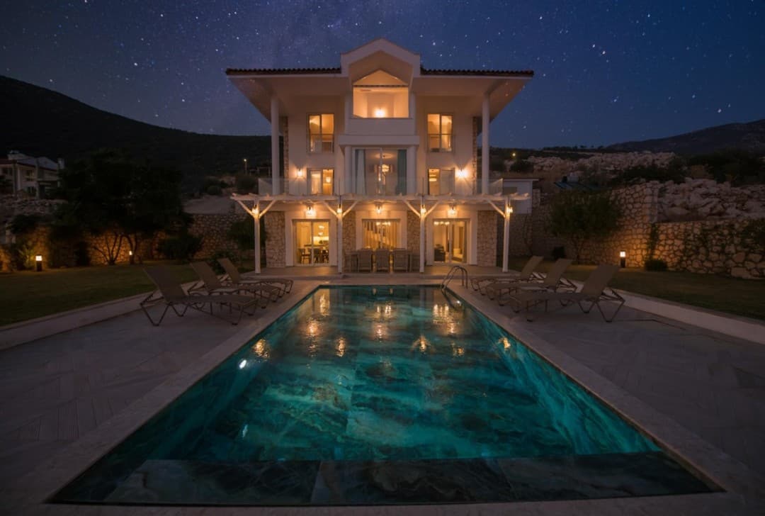 Gecenin karanlığında yıldızlı gökyüzü altında aydınlatılmış üç katlı villa ve önündeki açık havuz.