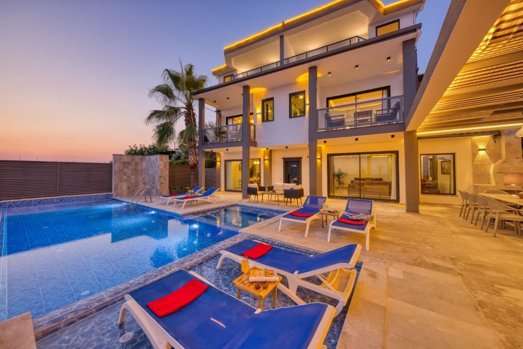 Gün batımında aydınlatılmış, havuz başında şezlonglar bulunan üç katlı modern villa.