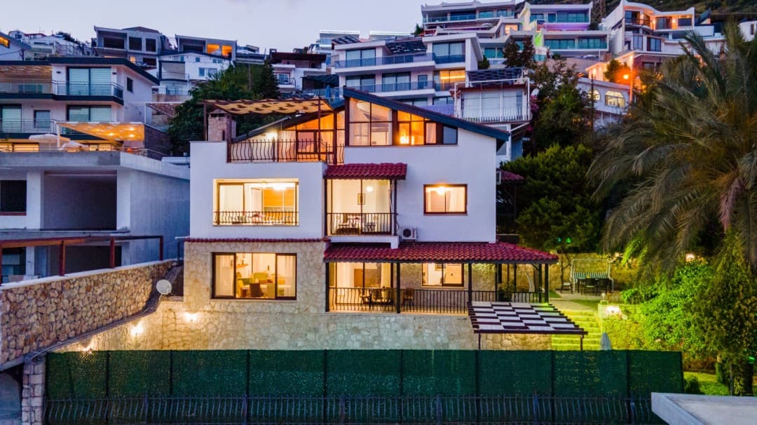 Akşam karanlığında ışıkları yanan, teraslı ve kiremit çatılı üç katlı villanın dış cephesi.