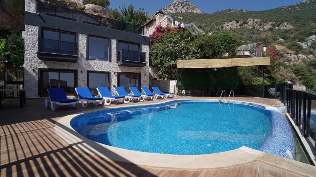 Ahşap şezlonglar ve mavi havuzun yer aldığı taş cepheli villa bahçesi.