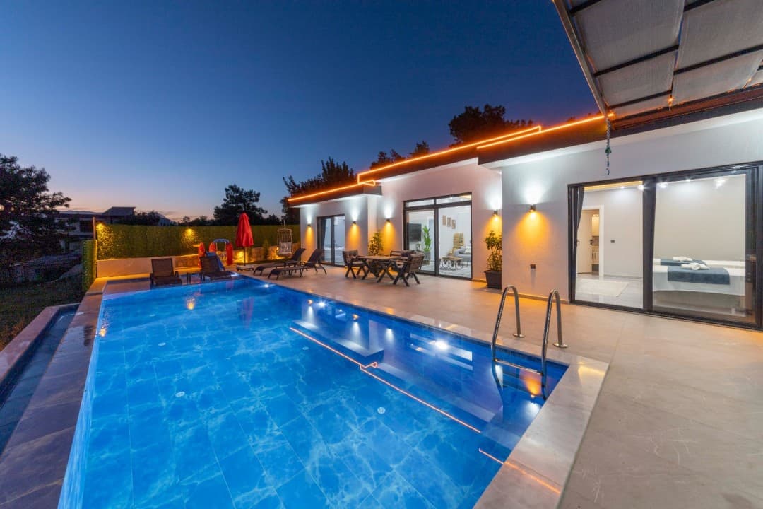 Akşam vakti aydınlatılmış havuzu, şezlongları ve dış oturma grubu bulunan modern tek katlı villa.