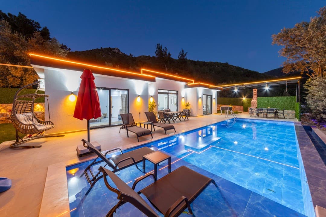 Havuz kenarında şezlonglar ve arka planda dağ manzaralı akşam aydınlatmalı villa.