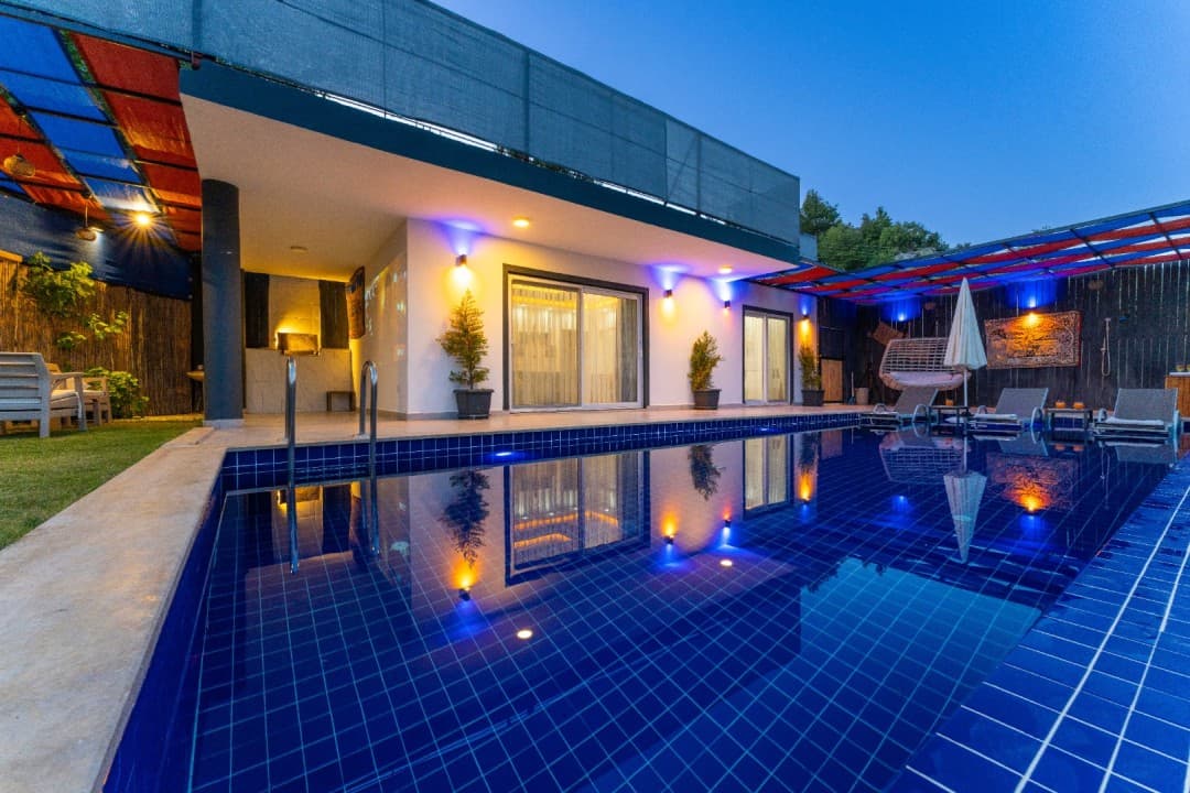 Akşam vakti aydınlatmalı havuz ve havuz kenarında şezlonglar bulunan modern villanın dış görünümü.