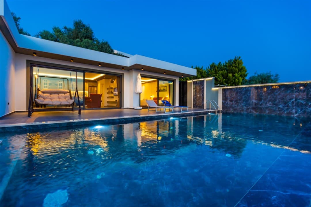 Akşam vakti aydınlatmalı özel havuzu, şezlongları ve bahçe salıncağı bulunan modern tek katlı villa.