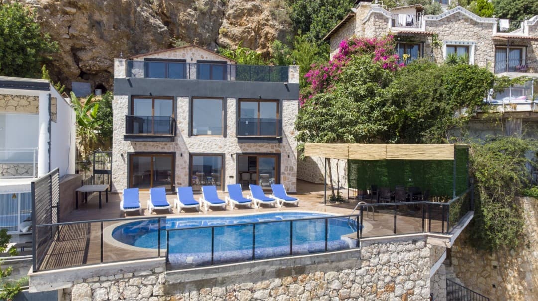 Kayalık dağ yamacında, önünde havuz ve şezlonglar bulunan çok katlı taş villa.