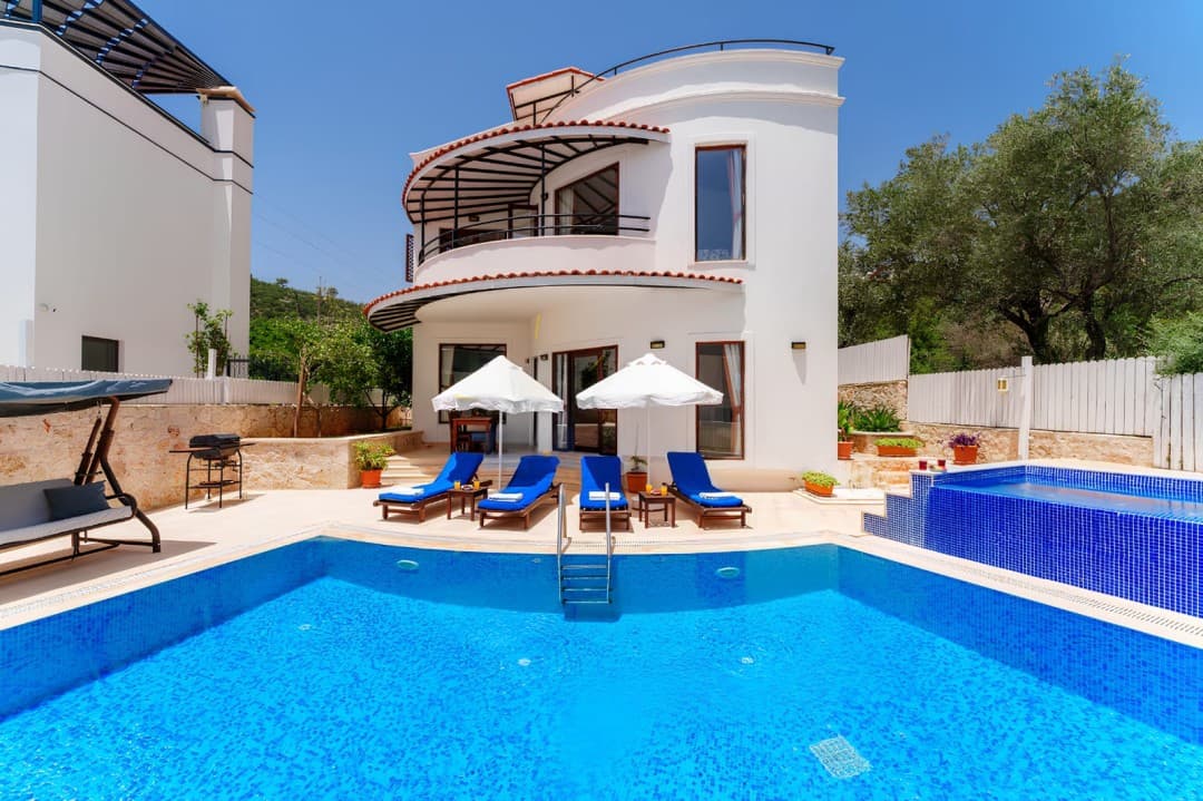 Önünde havuz, şezlonglar ve şemsiyeler bulunan iki katlı beyaz villa.
