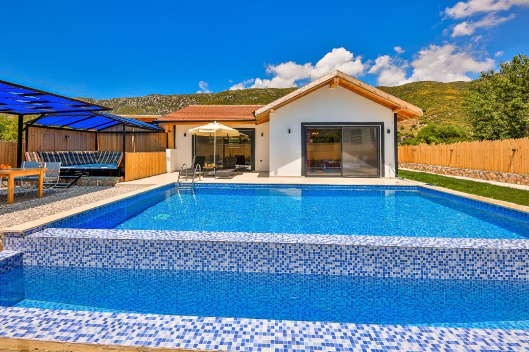 Mavi havuzlu, önünde güneş şemsiyesi ve oturma alanı bulunan beyaz dış cepheli müstakil villa.