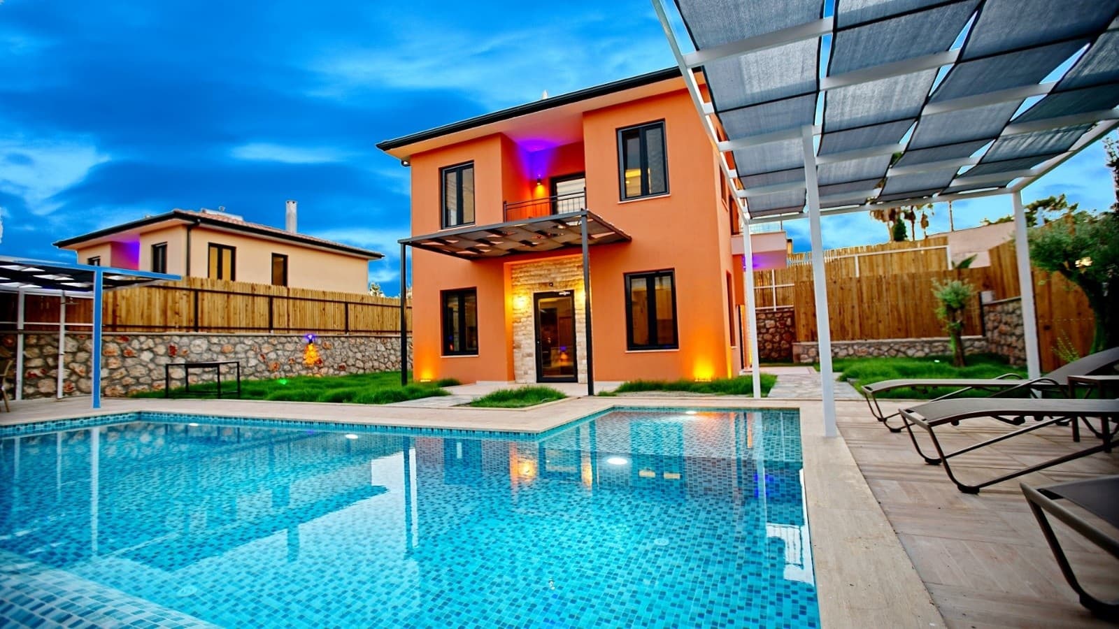 Önünde havuz ve şezlonglar bulunan turuncu dış cepheli iki katlı villa.