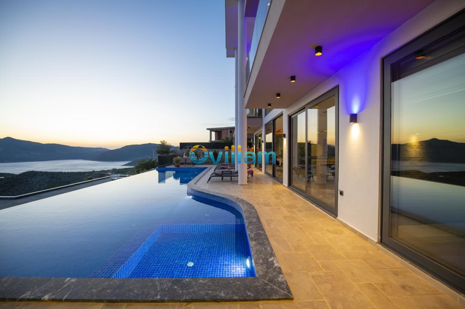 Deniz manzaralı havuz kenarında şezlonglar ve gün batımında aydınlatılmış villa dış cephesi.