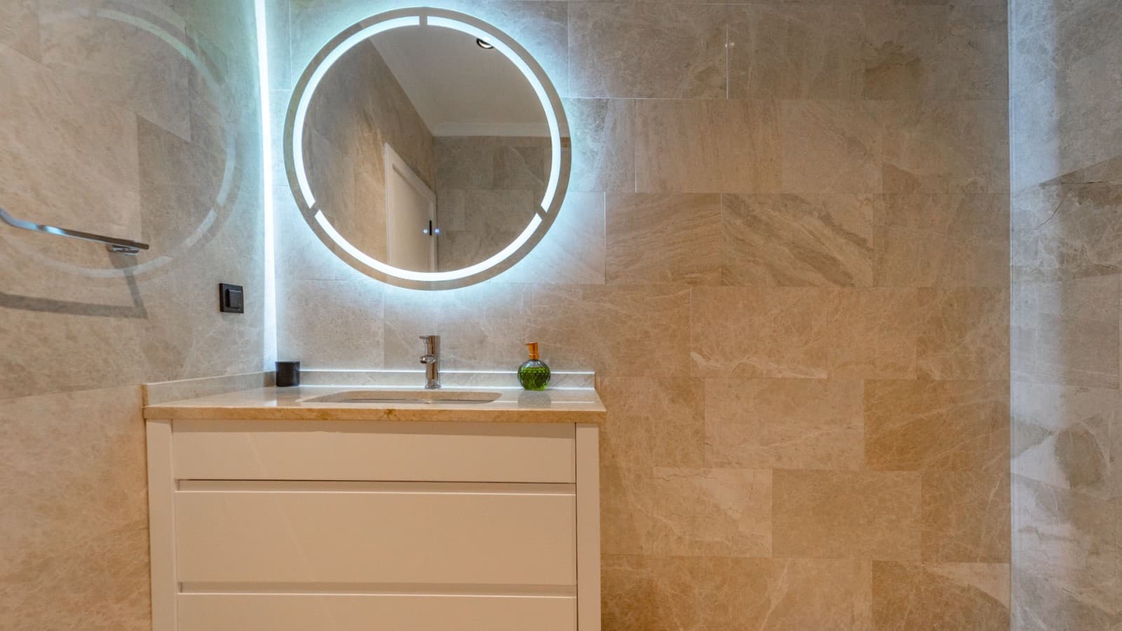 Bej seramik kaplı duvar önünde yuvarlak led ışıklı ayna ve lavabo.