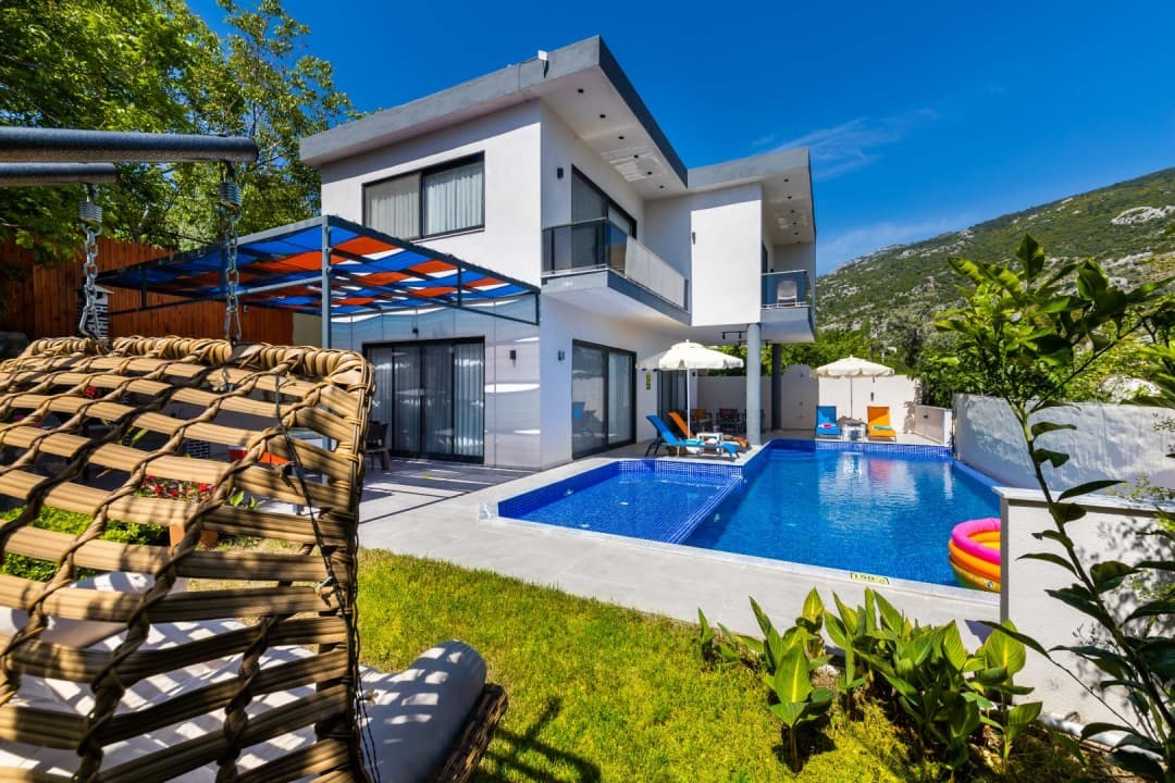 Ön planda hasır salıncak, arka planda havuzlu, beyaz dış cepheli iki katlı villa ve dağ manzarası.